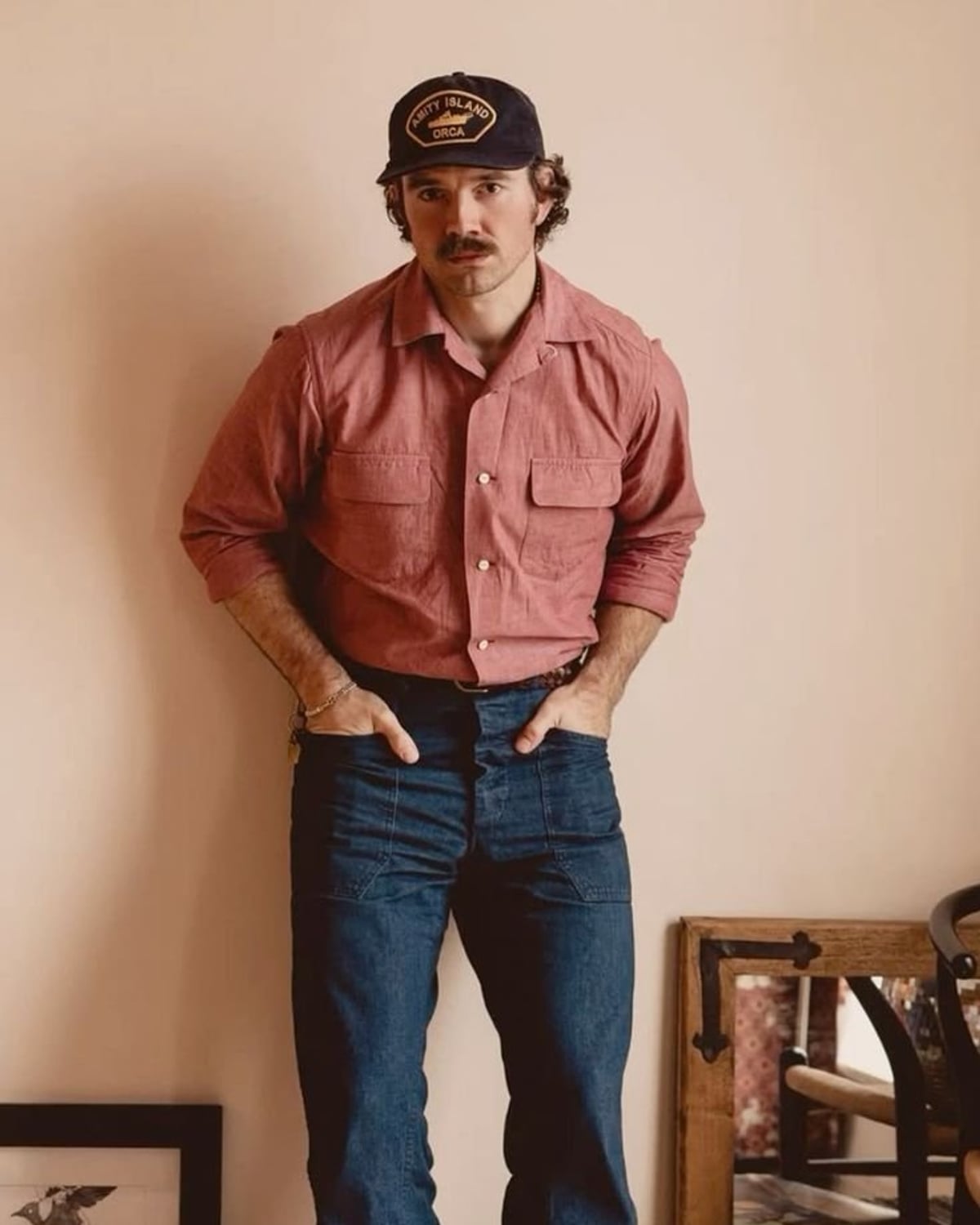 Brooklyn Vintage outfit — men's Amity Island Orca trucker cap, men's faded red chambray camp shirt, men's dark indigo bootcut jeans with patch pockets, men's gold chain bracelet, men's brown leather moc-toe boots