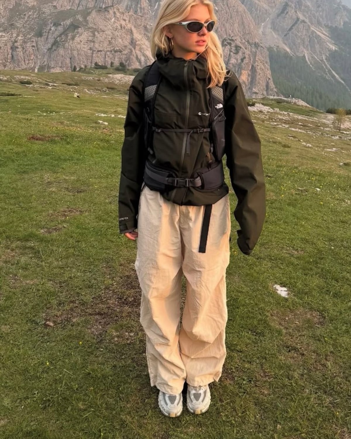 Gorpcore outfit — white wraparound sunglasses, olive GORE-TEX hooded shell jacket, black hydration vest backpack, cream parachute cargo pants, white chunky trail sneakers