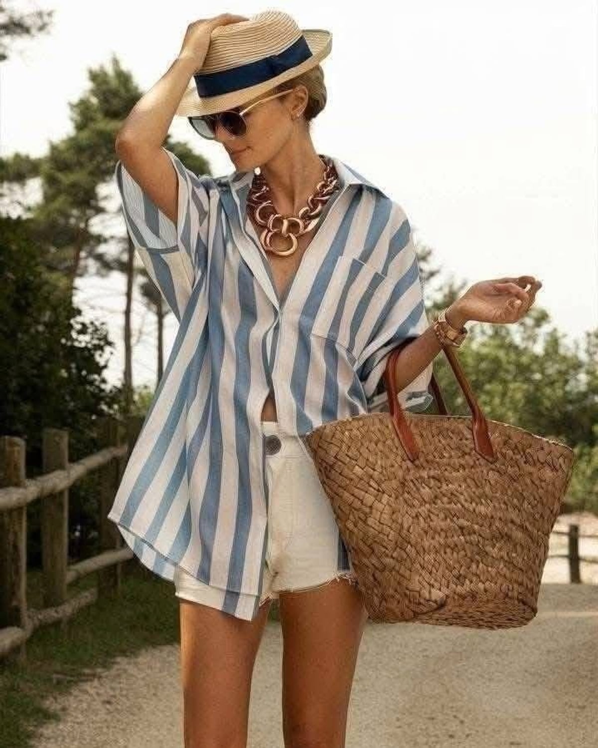 Riviera Resort outfit — straw sun hat with navy ribbon, tortoise oversized sunglasses, chunky gold link statement necklace, gold bangle bracelet stack, oversized blue-and-white striped button-down shirt