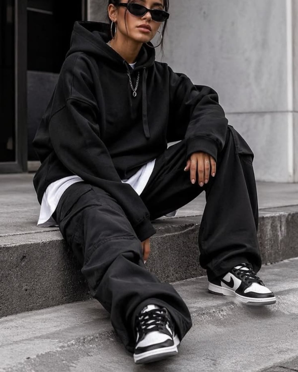 Streetwear outfit — black cat-eye sunglasses, silver hoop earrings, silver pendant chain necklace, oversized black hoodie, white layering tee